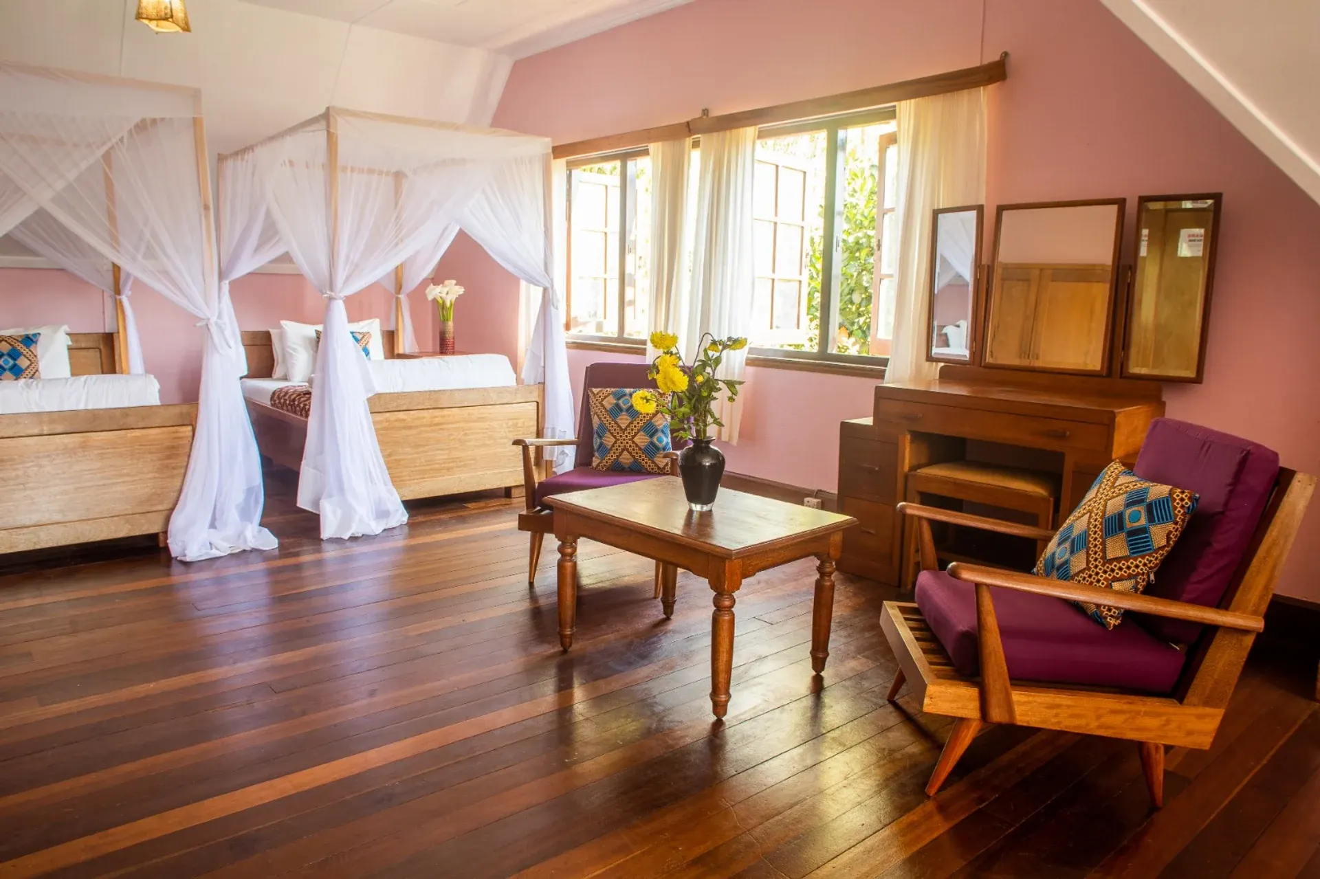 A pink-walled guest room at Mullers with twin beds under white mosquito-net canopies, a wooden vanity and armchairs on a polished hardwood floor
