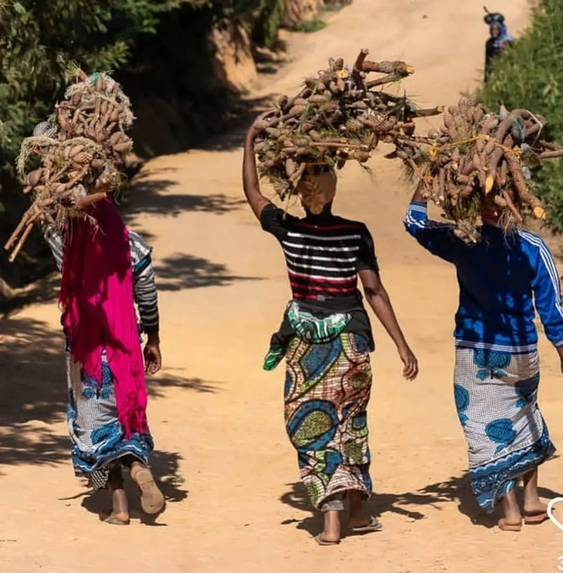 Three women in colourful kitenge wraps walking a dirt mountain road, carrying bundles on their heads near the lodge