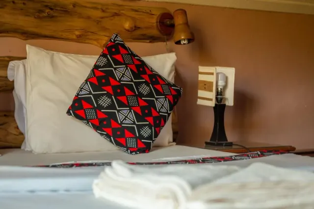 A made bed at Mullers dressed with a bold red-and-black kitenge cushion beneath a timber headboard and bedside lamp