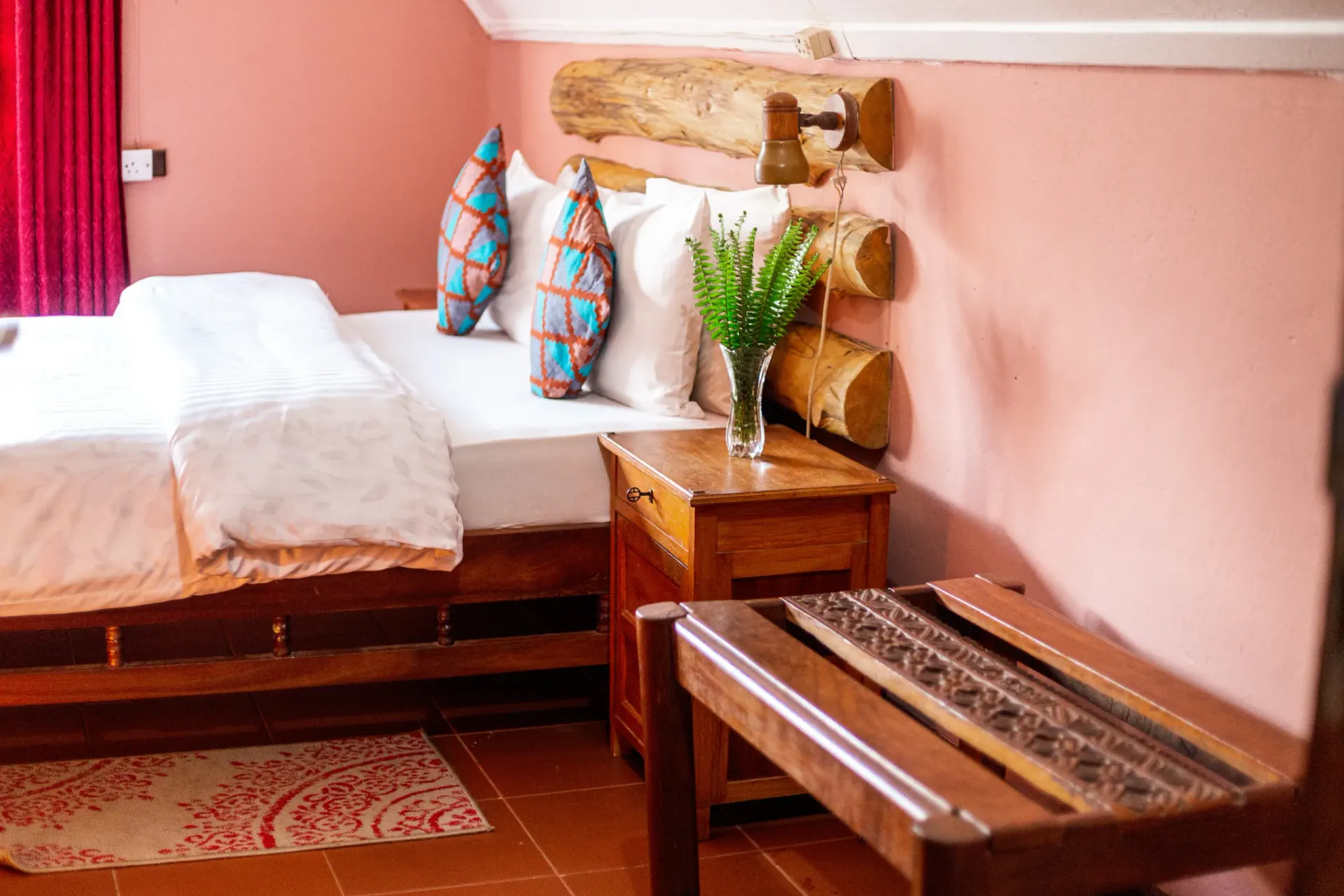 A main-house bedroom with soft pink walls, a timber-framed bed dressed in white linen, kitenge cushions and a carved wooden bench