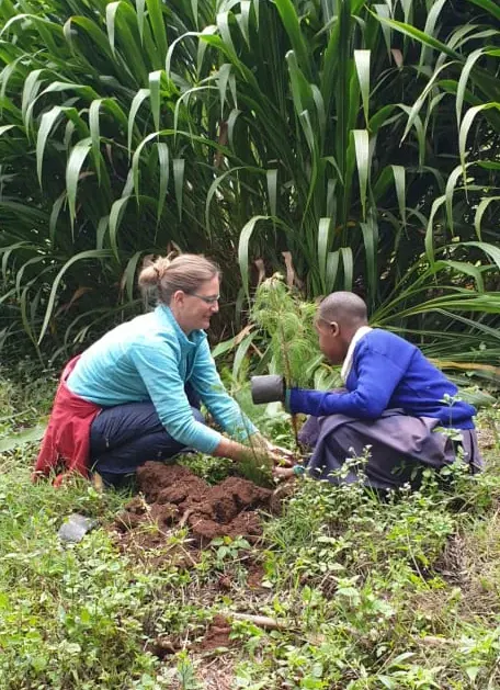 A guest and a local schoolchild kneel together to plant a tree seedling in the Usambara hillside