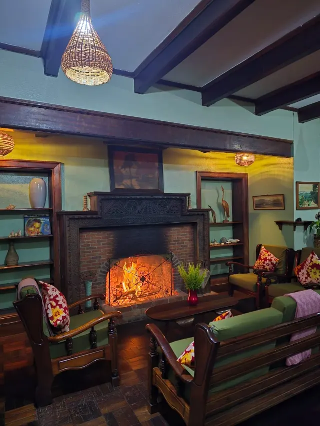 The lodge lounge with sage-green walls and a carved-wood fireplace lit at dusk, wooden shelves and floral-cushioned armchairs