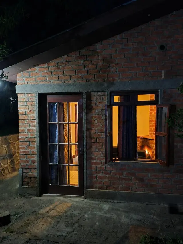 A brick guest cottage at night, warm light glowing from the door and an open window with a fire lit inside